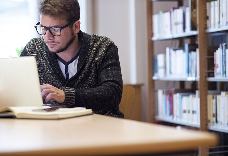 In einer Bibliothek arbeitet ein junger Mann konzentriert an seinem Laptop 
