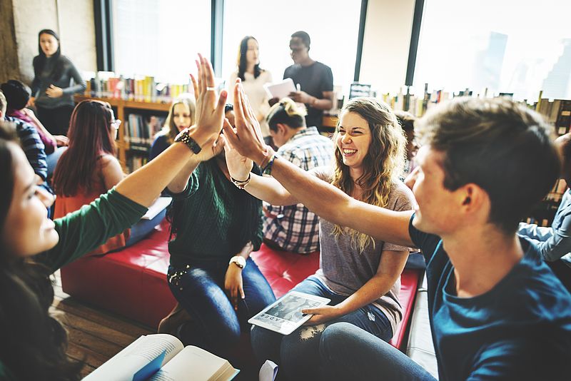 Viele junge Menschen sitzen zusammen, arbeiten und lernen, eine Gruppe hebt die Hände zum gemeinsamen High Five