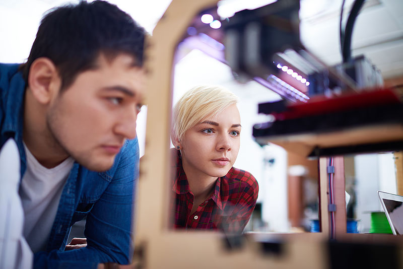 An einem 3-D-Drucker beobachten zwei junge Menschen den Druckvorgang